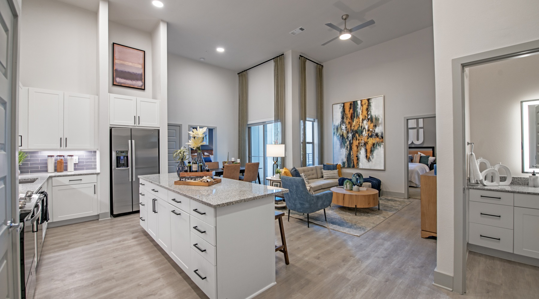 Spacious Living Modern open-concept apartment with a bright kitchen featuring white cabinets, stainless steel appliances, and a large island with seating. The living area includes tall windows, neutral flooring, and contemporary furniture with colorful accents.