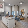 Modern open-concept apartment with a bright kitchen featuring white cabinets, stainless steel appliances, and a large island with seating. The living area includes tall windows, neutral flooring, and contemporary furniture with colorful accents.