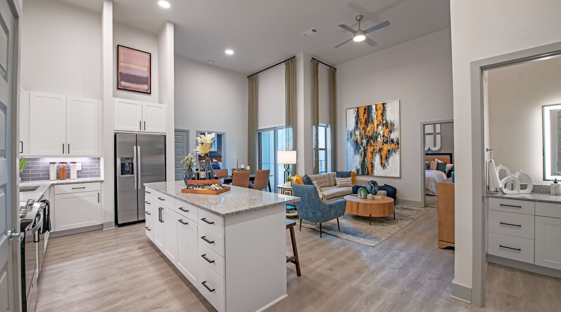 Modern open-concept apartment with a bright kitchen featuring white cabinets, stainless steel appliances, and a large island with seating at Alta at the Farm apartments in Allen, TX.