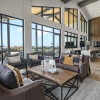 Spacious community lounge with floor-to-ceiling windows, stylish seating areas, wood-paneled accent wall, and a large TV. The room opens to an outdoor patio with views of the surrounding area.