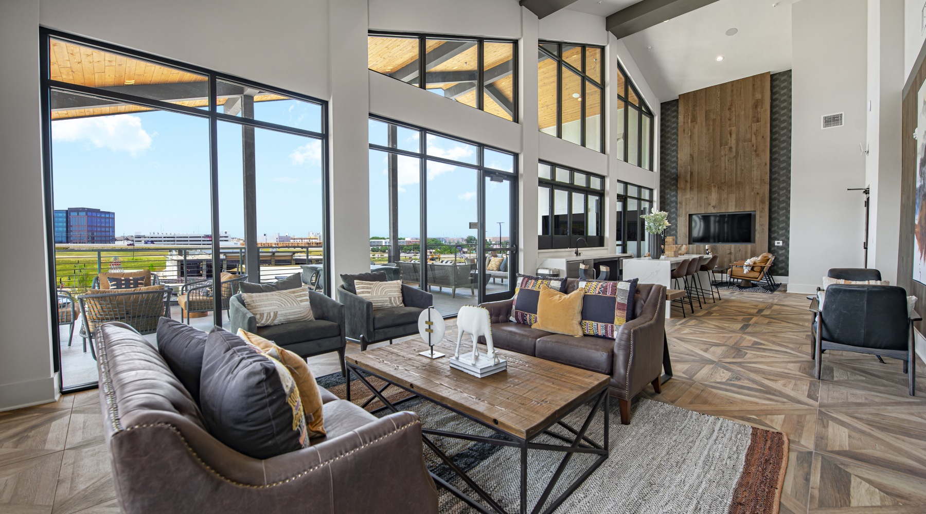 Sky Lounge Spacious community lounge with floor-to-ceiling windows, stylish seating areas, wood-paneled accent wall, and a large TV. The room opens to an outdoor patio with views of the surrounding area.
